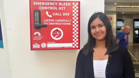 Sienna Ahir A girl with long dark hair, a silver necklace, white shirt and black jacket, smiles as she stands against a wall which has a red emergency bleed control kit mounted on it.