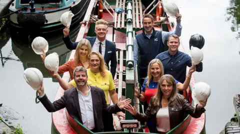 Eight people sit in a canal boat and hold up their own white builder's hat.