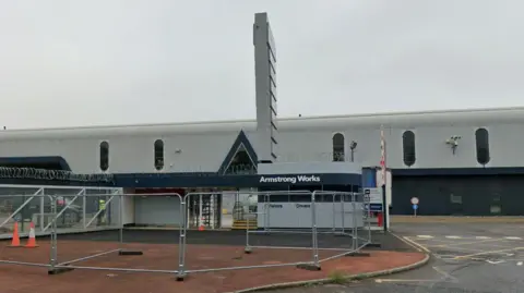 The Armstrong Works site on Newcastle's Scotswood Road. A gatehouse is located in front of the building which is made from grey corrugated metal.