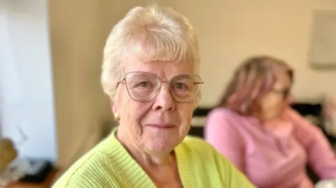 Martin Giles/BBC Sandra Taylor is sitting at a table in the community center wearing a bright lime-green knitted sweater and glasses. Sandra is smiling at the camera. In the background, another person is blurred but you can see she is wearing a pink top and has shoulder-length hair with reddish tones. 