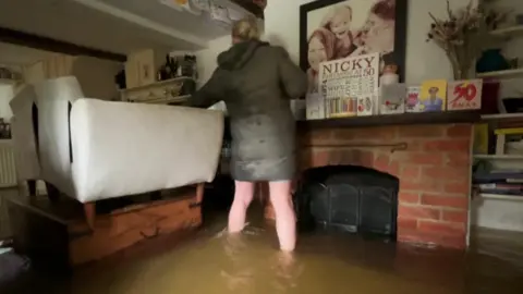 A woman stands in her living room. There is brown water up to her shins. A sofa is balanced on top of a wooden coffee table, out of the water. There is also a brick fireplace with family pictures and birthday cards on top.