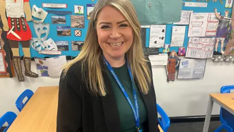 Sarah Brinicombe/BBC Mary Gates, the headteacher at Edenbridge Primary School, pictured in a classroom at the school. She is stood in front of a wall with children's drawings and paintings. Mary is wearing a green dress, black jacket and blue lanyard. She has long blonde/brown hair.