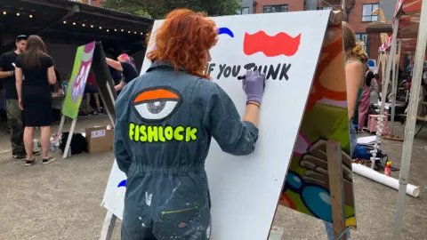 An artist paints on a canvas outside wearing a set of blue overalls with the word fishlock written on the back in yellow letters under a painting of an eye. There are other artists working on pieces around her.