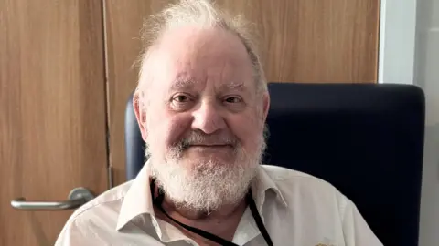 Royal Papworth Hospital Roderick Starksfield sits inside his room while at Royal Papworth. He is wearing a cream shirt and has a grey beard and grey hair. He has a black lanyard around his neck.