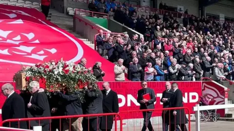 A group of men carry Phil Lowe's coffin which is covered in flowers. Crowds of people in the stands clap in the background.