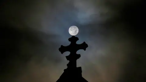 Lucie Johnson A nighttime shot of the top of an apparent church building in shadow with the moon in the sky above. The sky is mainly black but there is a slight white glow around the moon. The top of the church is in the shape of a cross which is in darkness.