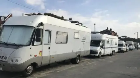 A row of light coloured campervans, parked up bumper to bumper on a road. Above can be glimpsed roofs of buildings. In front is a wide tarmacked road or pavement. 