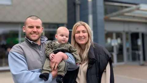 The University of Southampton A family of three pictured outside. The dad is to the left wearing a light blue and grey fleece while holding a baby which is wearing a khaki tracksuit. The mum has long blonde hair and is wearing a beige jumper with a black gilet over the top.