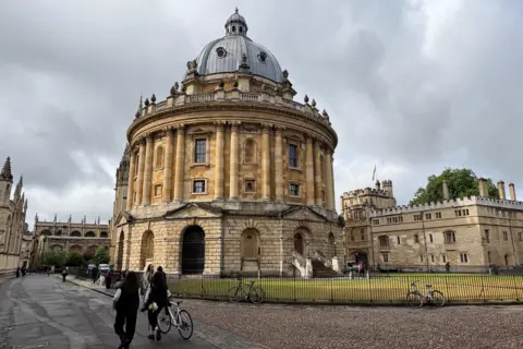 AngelaJ The Radcliffe Camera is a round stone building. In front is a grassy area and there are buildings surrounding the square. There are bikes resting against the black railings and two people are walking past pushing a bike.