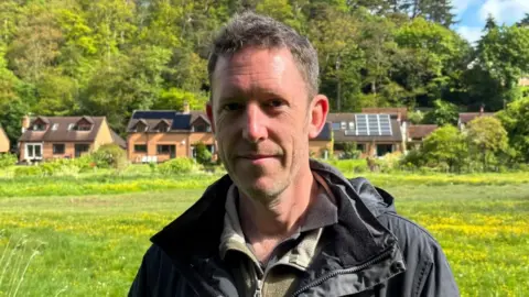 There is a man with short brown hair. He has stubbly facial hair and a slight smile. He is wearing a black coat, a green jacket and a brown t-shirt. Behind him is a big green meadow, with a row of houses behind him, 