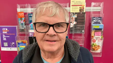 A man wearing a black jumper and black jacket, also wearing glasses, is stood in front of a red wall which has a display behind it - the display is containing a variety of leaflets. 
