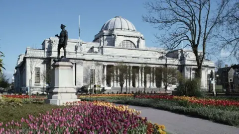 The Edwardian National Museum building in Cardiff's Cathays Park, with a white dome, and a statue and a flower garden in the foreground and a flower garden