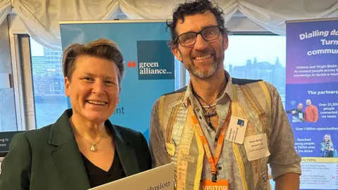 Sarah Dyke Sarah Dyke is standing next to Simon Barfoot in a room with posters behind them. They are both smiling at the camera.