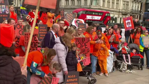 A protest in central London. Participants are in orange and holding placards. A red double-decker bus is in the background