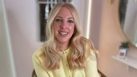 Laura Rush has long wavy blonde hair. She is smiling, sitting in a hairdressers chair and wearing a light yellow cardigan.