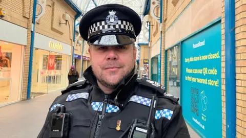 A policeman wearing his uniform with radio and body camera attached. He is wearing his police hat. He is standing in a shopping centre.