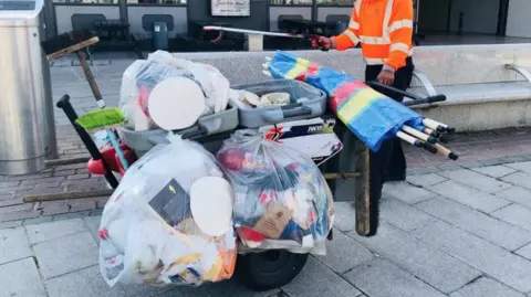 A man wearing orange high-vis collects waste on a cart on a street, with in bags full of rubbish.