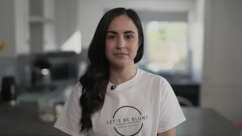 Leanne Lucas, who has long wavy dark hair, and is wearing a white t-shirt with a circular logo with the catchphrase 'Let's Be Blunt' on the front. She is standing in a kitchen looking into the camera with a determined expression.