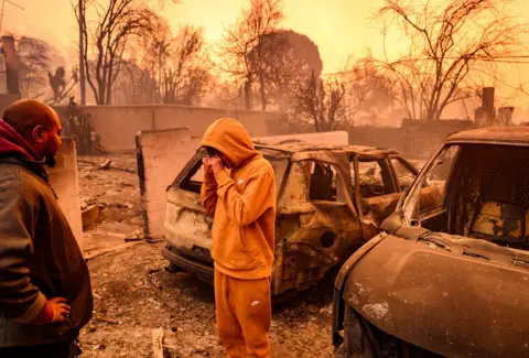 Getty Images Seorang pemuda menangis saat ia mensurvei reruntuhan sebuah mobil yang terbakar di kebakaran Los Angeles. Pria lain melihat.