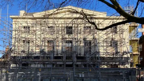 A wide three storey building, cream coloured, with large windows. Scaffolding is wrapped around it.