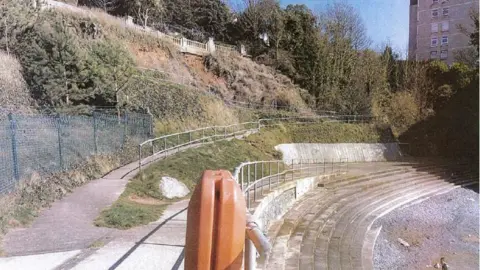 Kay Elliot A side view of Beacon Cove. There are steps at the top leading down to a rocky beach. On the pathway above, there is a pathway, with a patch of grass in-between. At the front, there's an orange safety float attached to the silver railings surrounding the beach area. 