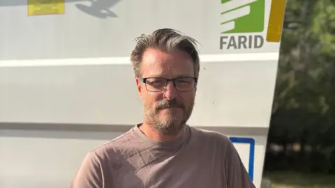 BBC A man wearing glasses and with a greying beard smiles at the camera while standing in front of a recycling truck bearing the word "Farid".