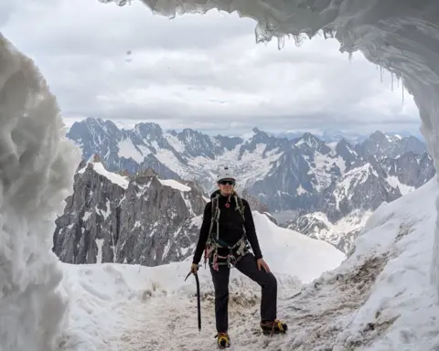 @el.ventures.earth A photo of a woman in black clothing and hiking gear, with a helmet and sunglasses, stood on the snow in front of snow-topped mountains