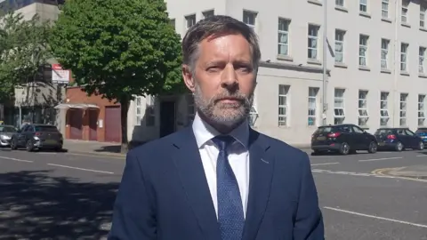 A man with short grey hair and a grey beard, wearing a blue suit jacket and tie with white shirt, he's stood on a street with a large white building behind him, several parked black cars and a large tree