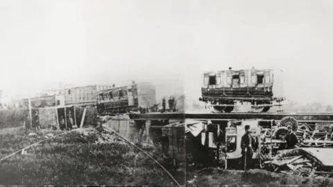 Getty Images Old train carriages on a viaduct, some partially off the rails. Other carriages are destroyed underneath the viaduct in the black and white image.