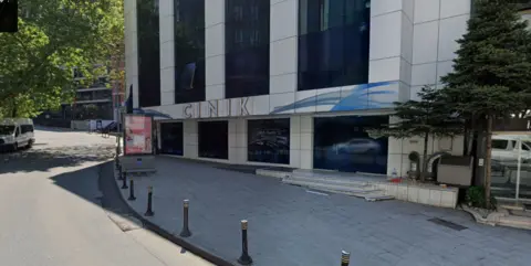 Google A modern commercial building with a white exterior and large dark‑tinted windows beside a curving urban street. The building has metallic letters spelling CINIK above the doorway. Short black bollards line the pavement in front.