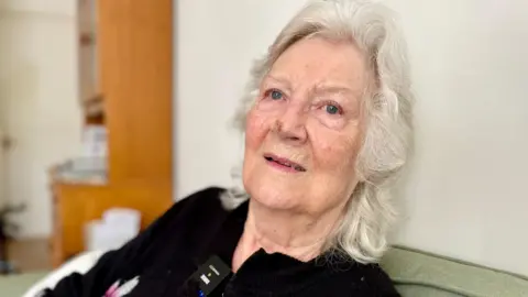 Mark Norman/BBC A woman with grey hair wearing a black blouse witting in a living room. She is leaning back against a green sofa cushion and in the background there is a large wooden shelving unit.