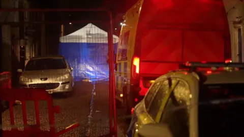 A blue tent with a white roof lit up by car lights. A marked police van is parked facing away from the camera. A market police car is parked behind it facing the camera. There is also a silver car parked close to the tent and facing the camera. Metal gates surround the scene. 