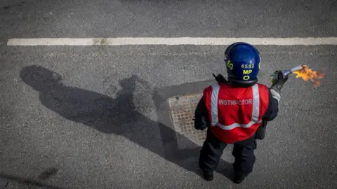A man in a blue helmet and a red high-vis jacket labelled 'instructor' carrying a lit petrol bomb in his right hand.