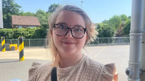 Hannah Gray/BBC A young woman with tied-up blonde hair in a beige shirt. She is wearing black and brown glasses and there is a car park behind her.