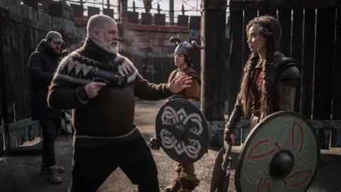 Universal Pictures A white haired bearded man gives direction to an actor. He wears a brown and cream nordic knit, holds a camera in his hand and gestures with the other hand. The actor he gestures to is a girl with long brown hair, dressed in viking warrior clothing and she holds a round colourful shield