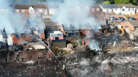 An aerial shot of a row of homes with their roofs destroyed by fire. Smoke streams into the air. Land in front of the houses is scorched, with firefighters holding hoses spraying water towards the scene.