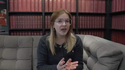 Erin, who has long dark blonde hair and glasses, sits on a grey sofa with a bookcase in the background, speaking to the camera