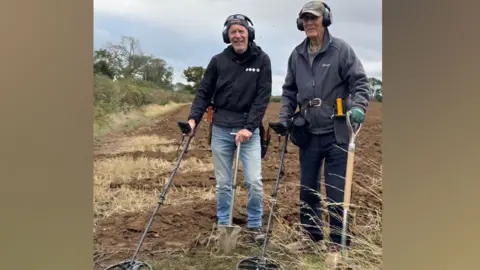 Peter Derby Dave Derby and Peter Derby in a ploughed field in September 2025. Peter is on the left of the frame, dressed in jacket and jeans, and Dave on the left, dressed in a fleece and trousers. Both are wearing headphones over their ears and have metal detectors in their right hands and spades in their left.