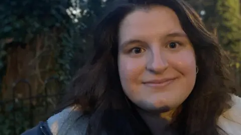 Woman looking straight at the camera slightly smiling, she has dark brown hair and brown eyes. She is wearing a blue coat with cream fluffy lining