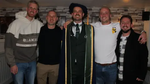 Abbie Garcia Matty Lewis, Dan Braidley, Jack Woodhams, Mike Hewitt and Sam Watling stand shoulder to shoulder. Jack, in the middle, wears a graduation cap and gown.