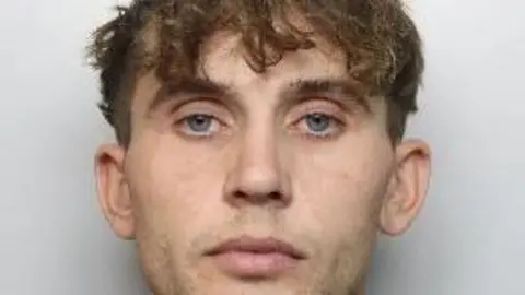 A mugshot of a man who looks directly at the camera. He has brown hair, slightly curly, which covers his forehead.