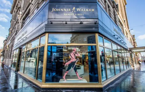 Getty Images The front entrance to the Johnnie Walker Experience in Edinburgh has a window with a print of a man striding in a red coat, tall boots and hat with a red cane and black trousers. A black panel is above the window and reaches around the corner. It says "Johnnie Walker, Princes Street"