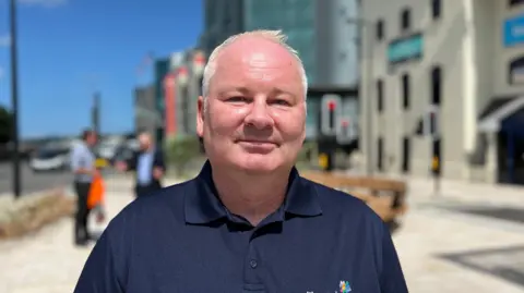 A person wearing a navy polo shirt with a LibertyBus logo stands on a wide city pavement. Modern office buildings and a few pedestrians appear blurred in the background under a bright blue sky with scattered clouds.