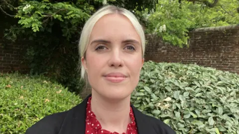 Gemma has blonde hair and brown eyes. She is wearing a black jacket and red top with white polka dots. Her hair is tied back behind her head. She is looking down the camera lense and is stood in front of some green bushes.