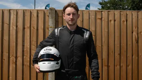 Eastern Education Group Brady Pollock standing in front of a brown fence, dressed in all-black race gear whilst holding his white crash helmet.