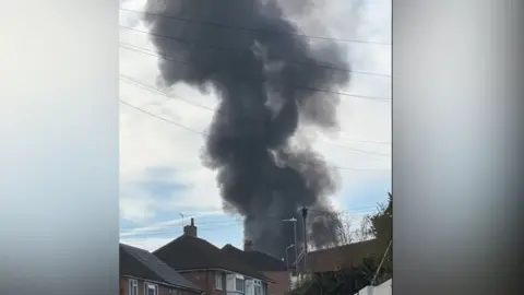 A big black plume of smoke pumps into the sky above the roofs of houses and electricity wires