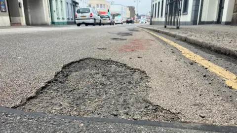 BBC A pot hole on a main road with traffic on the left in the background.