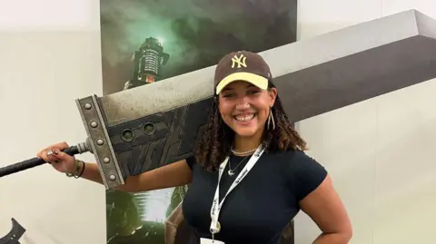 Imogen Mellor Imogen is wearing a black t-shirt and brown and yellow cap. She smiling at the camera holding a wide sword prop over her shoulder. She has brown curly hair and is wearing a pearl necklace and dangly earrings. 