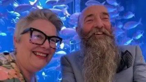 Handout John Gifford and Lisa Gifford pictured smiling in a blue lit room. John has a long grey beard and a moustache which curls at the ends, he wears a blue suit jacket with a dark blue pocket square while Lisa has a grey pixie cute, wears red lipstick, large black framed glasses and a pink, yellow and blue floral top.