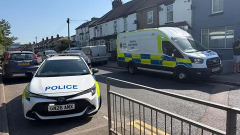 Swanscombe Neighbourhood Watch Two emergency vehicles parked up on a residential road. One is a police car the other is a police van. There are other cars on the road too and a row of terraced houses. 
